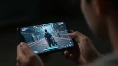 Um jogador concentrado olhando para a tela de um smartphone, que exibe uma cena de ação intensa de um jogo com gráficos realistas, em um ambiente escuro.