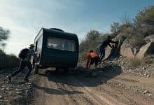 Um trailer recreativo atravessando uma estrada acidentada em meio a uma paisagem selvagem, com amigos trabalhando juntos para seguir viagem