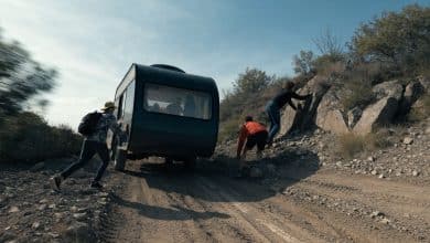 Um trailer recreativo atravessando uma estrada acidentada em meio a uma paisagem selvagem, com amigos trabalhando juntos para seguir viagem